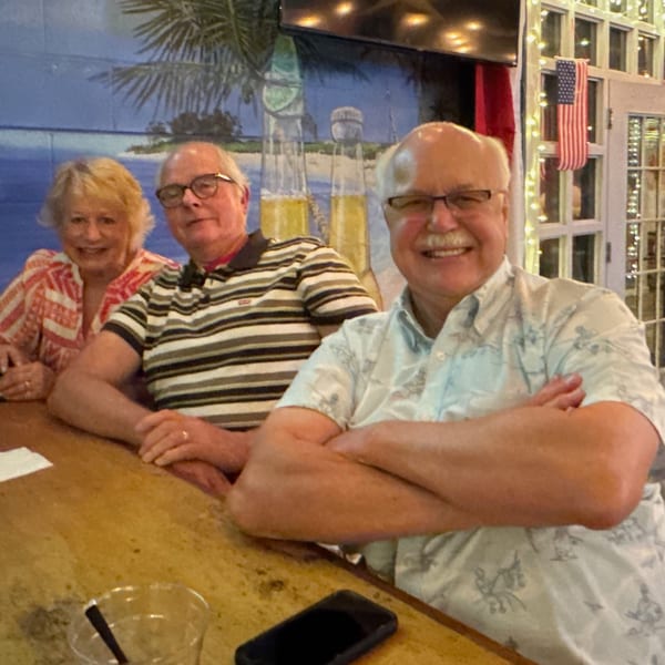 Three smiling people sitting at a bar with a tropical mural and American flag in the background.