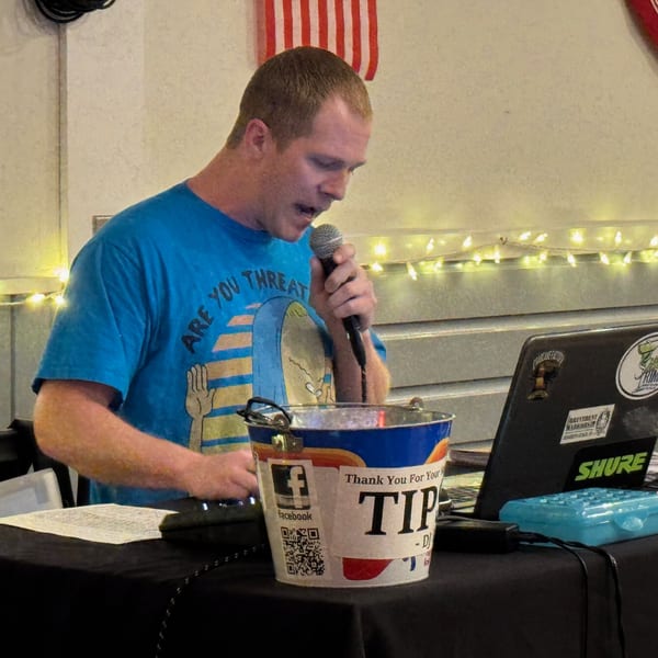 A man in a blue t-shirt sings into a microphone next to a tip bucket and laptop.