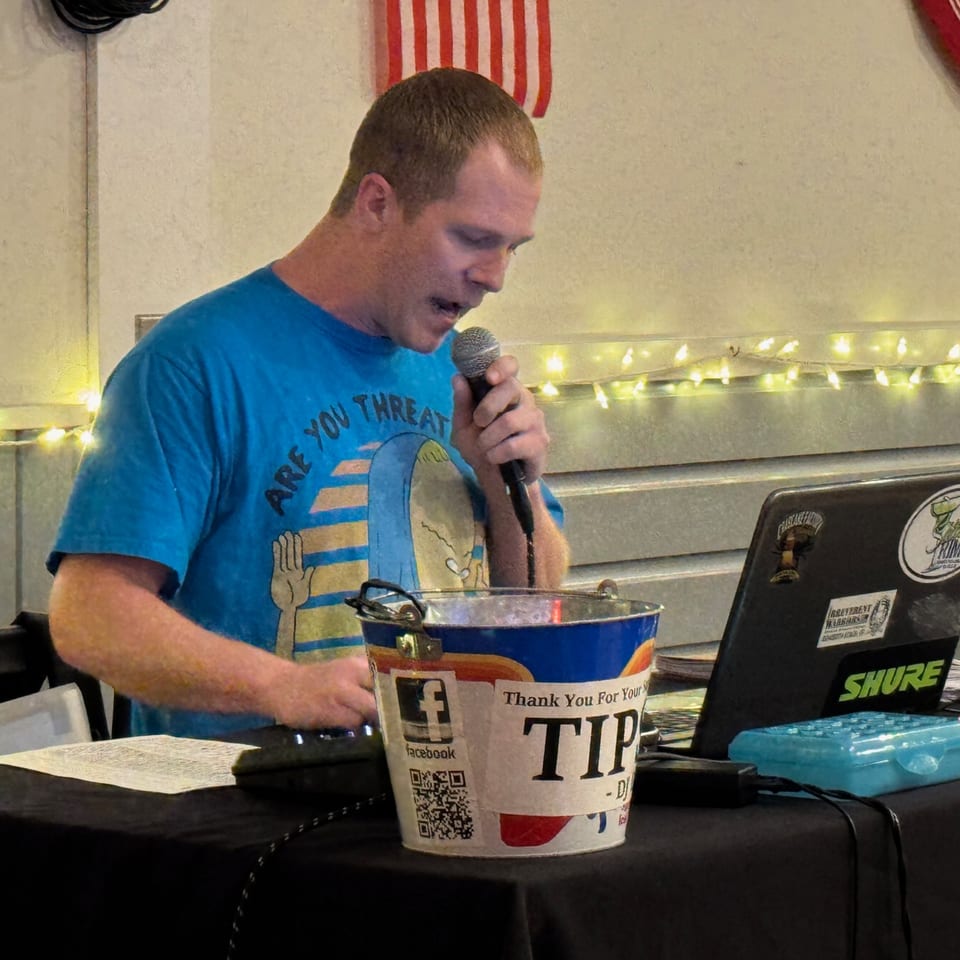 A man in a blue t-shirt sings into a microphone next to a tip bucket and laptop.