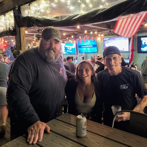 Three smiling people pose for a photo at a lively bar with string lights and American flags.