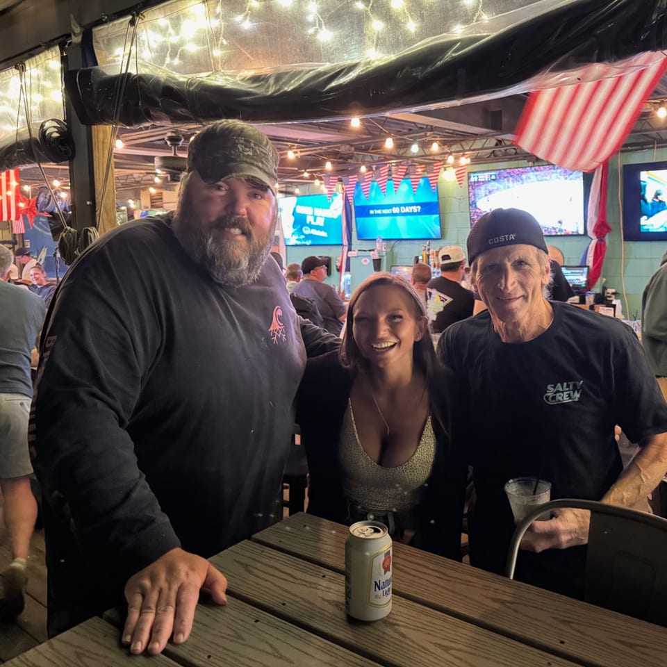 Three smiling people pose for a photo at a lively bar with string lights and American flags.