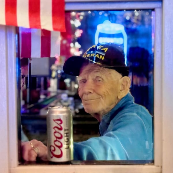 An older man in a blue jacket and veteran hat smiles, holding a Coors Light can.