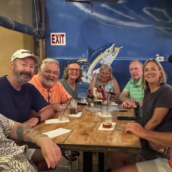 Six happy customers seated around a wooden table with drinks, against a blue wall with a fish mural.