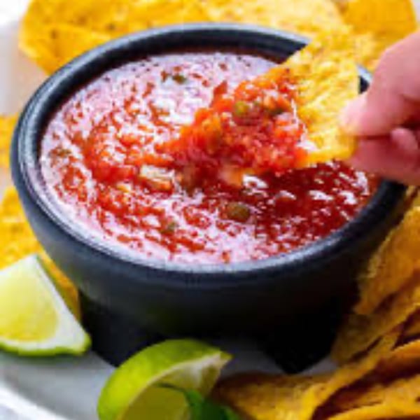 A hand dips a tortilla chip into a black bowl of vibrant red salsa, with lime wedges nearby.